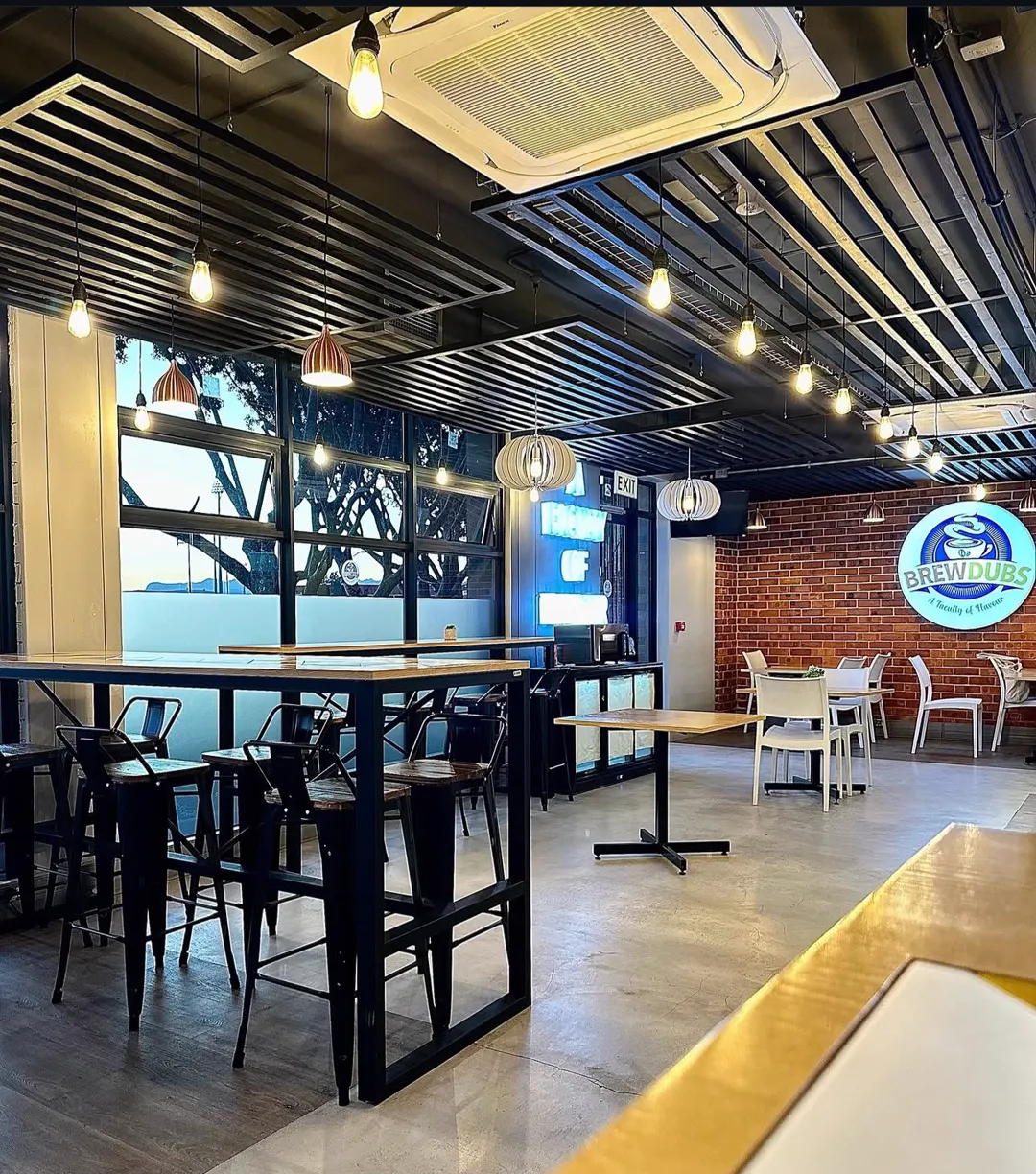 High bar tables with black stools along a window, low tables at the back, and the logo lit on a red brick wall.
